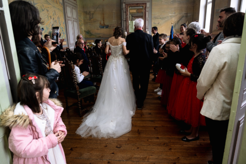 Mariée s'avançant devant le bureau de la mairie au bras de son père. Photo prise de dos dans un style pictural